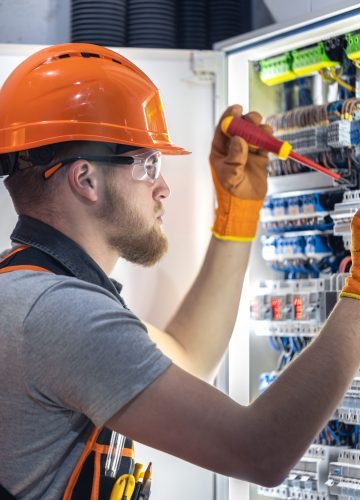 Male electrician working in switchboard. Male electrician in overalls working with electricity. High quality photo. Close-up. Man wearing safety glasses and hard hat.